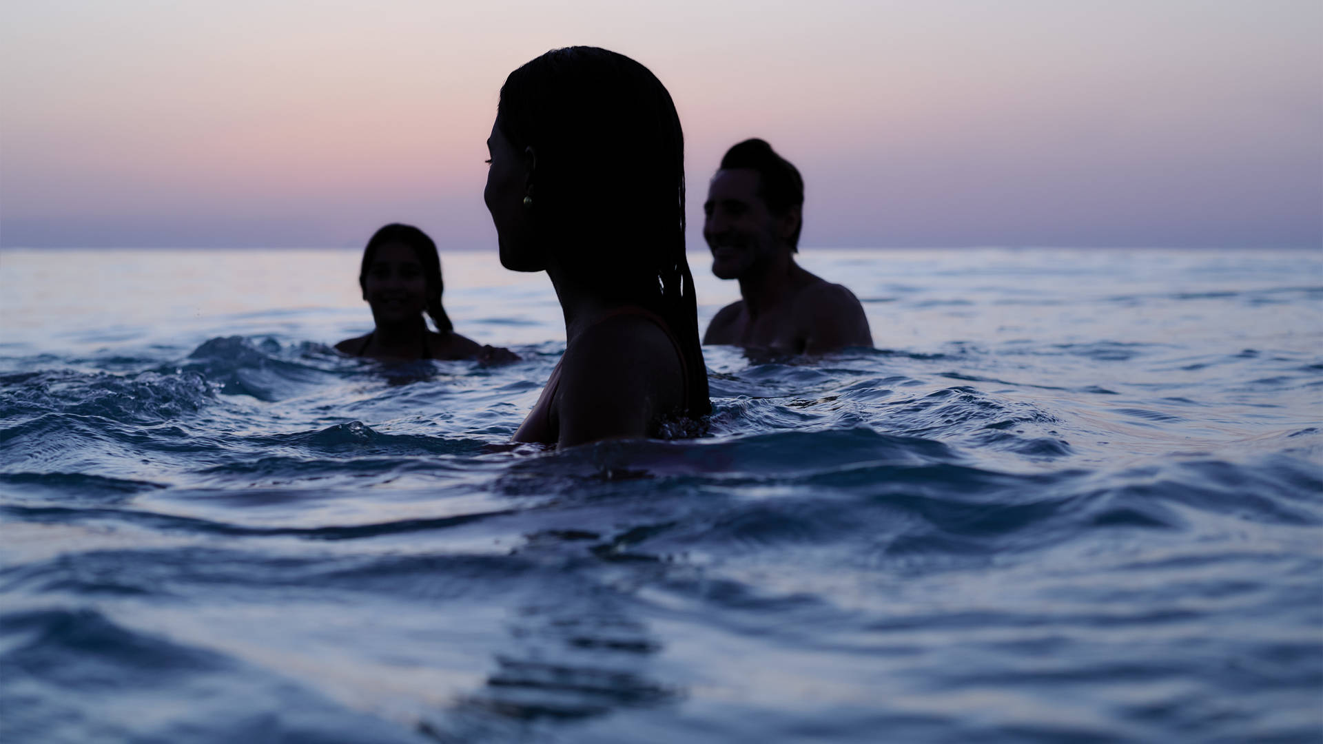 A shadowy outline of three people swimming in the ocean at twilight, the purple horizon behind them