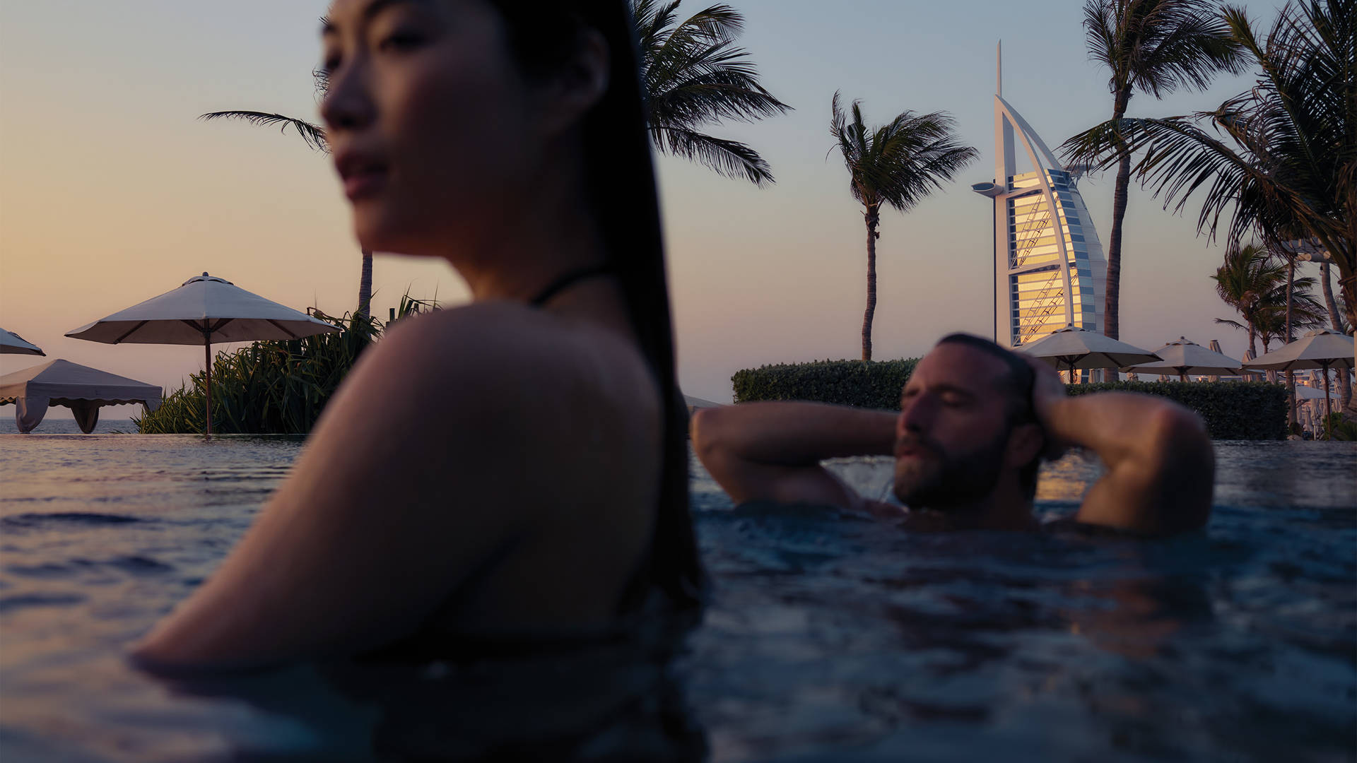 Image of a man and woman relaxing in the swimming pool at sunset, with the Burj Al Arab in the background