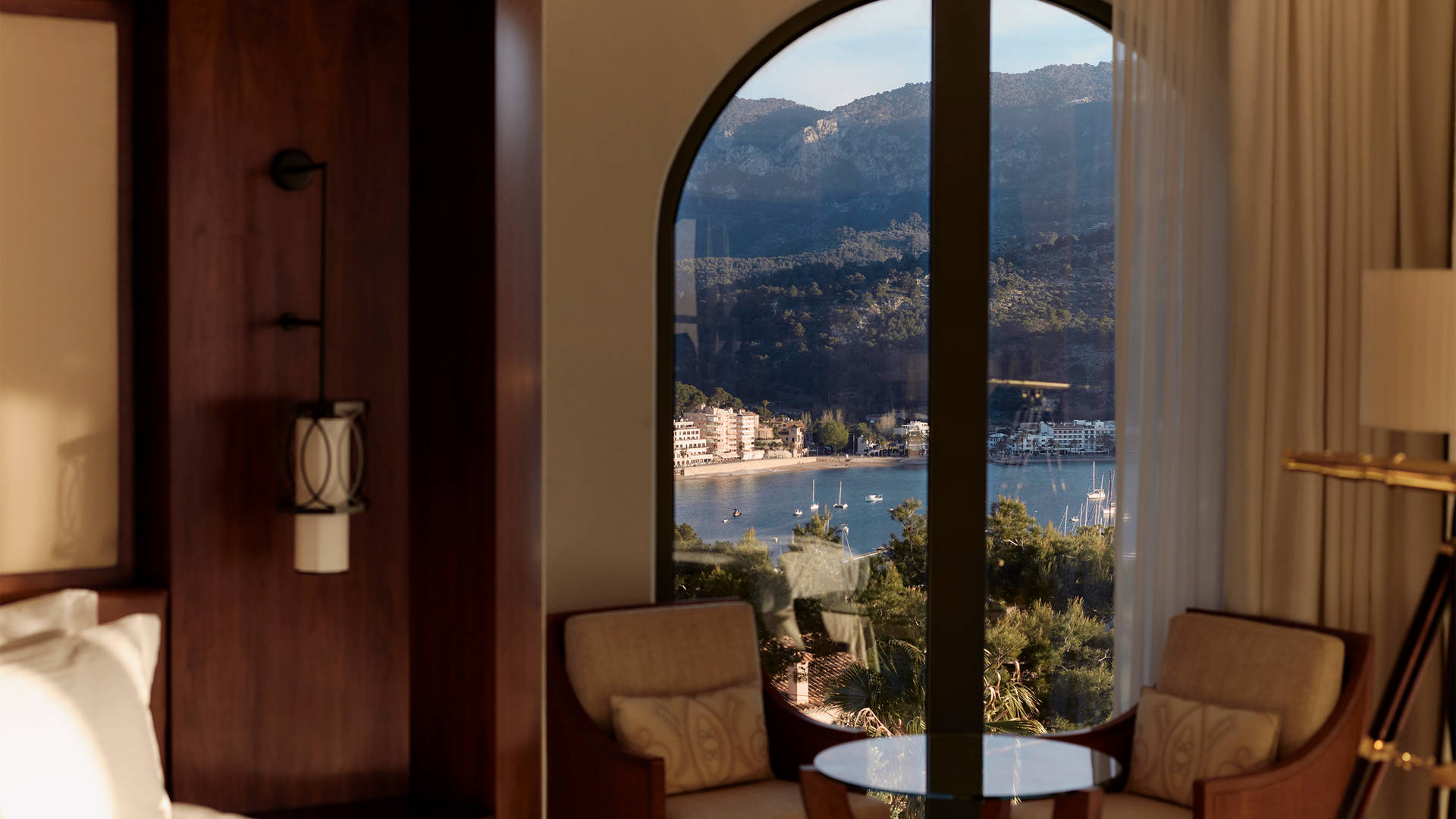 Image of a sunlit hotel room at Jumeirah Mallorca, with a window overlooking the sea and surrounding mountains