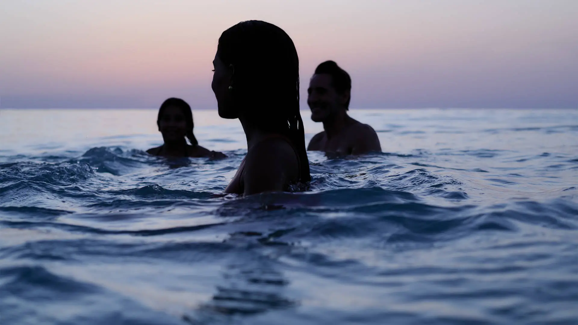 A shadowy outline of three people swimming in the ocean at twilight, the purple horizon behind them